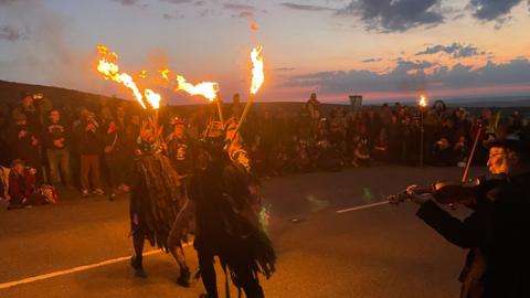 A picture of the Morris dancers dancing with fire on Haytor. They are wearing a black outfit and dancing in the road. Crowds can be seen around them.