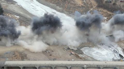 Dark and light grey smoke emanates from a shrouded bridge next to a dam.