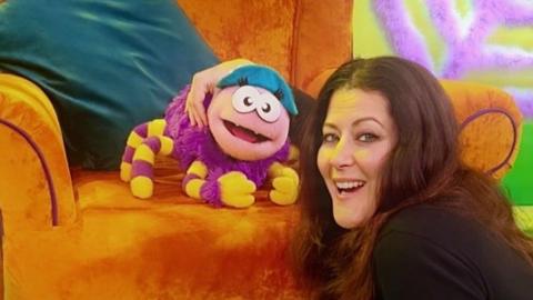 A female puppeteer smiles at the camera with one of her puppets, a purple spider