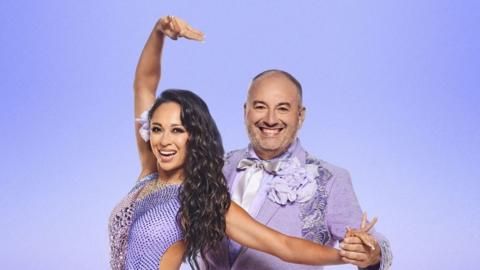 Studio portrait of Katya Jones and  Wynne Evans posing together against a plain blue background. They are looking at the camera and smiling and  Evans is holding Katya Jones's hand.