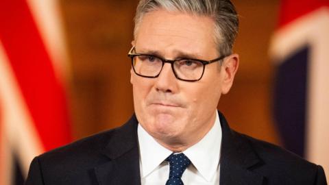 Keir Starmer in a black suit, white shirt and blue spotted tie has a solemn look on his face as he stands in front of union jacks in a room of Number 10, Downing Street, which are blurred in the background.