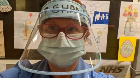 A woman in blue medical scrubs is taking a selfie. She is wearing red glasses, with a large, clear, medical visor, which covers her entire face. Underneath, she is wearing a face mask over her nose and mouth.