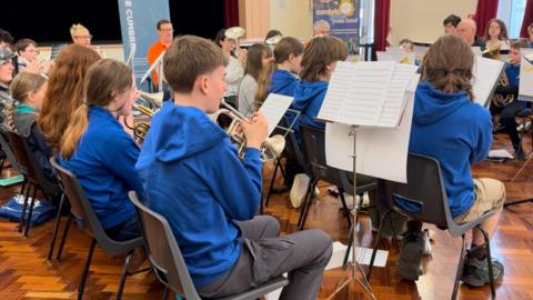 Young musicians wearing blue hoodies play brass instruments in a rehearsal room. They are sitting on grey, plastic chairs on parquet wooden flooring and are following sheet music.