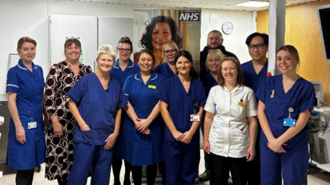 A group of people dressed in blue or white nursing scrubs posing for a picture. In the background there is a poster of a woman with the NHS logo at the top right.