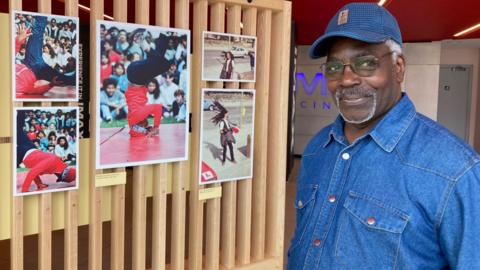 A man wearing a blue denim shirt and blue cap standing next to colour photographs on a wooden slatted display wall. 
