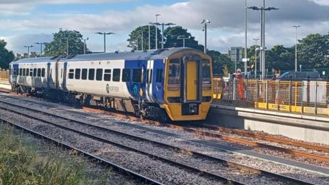 A blue and yellow Northern train at Ashington Station. It is a sunny day casting shadows across the three tracks. There is a workman in a hard hat and orange overalls leaning on railings on the platform.