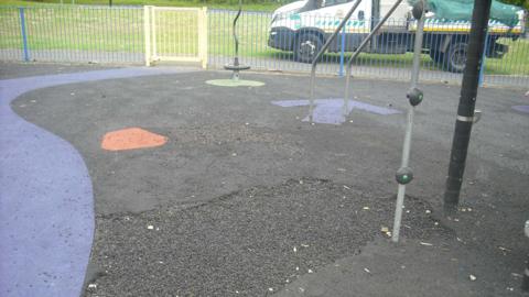 Black rubber surfacing with blue and orange spots next to play equipment. A large hole is in the floor. 