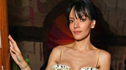Lily Allen, with her dark hair tied up in a bun, stands with her hand against the wall and looks at the camera. She is wearing a low-cut gold dress with big sequins