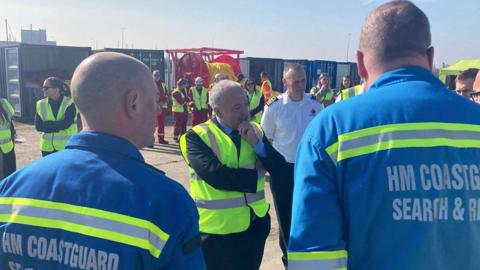 Transport Minister Mike Kane stands with his arms folded, wearing a dark suit and high visibility vest, talking to HM Coastguard search-and-rescue workers who have their backs to the camera and are wearing blue overalls 