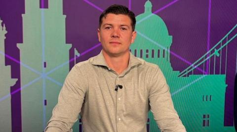 A man with short dark hair and a grey shirt smiles for the camera. A screen is behind him showing silhouettes of buildings. 