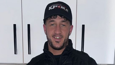 A photograph of Kaine Fletcher, a mixed-race man in his 20s, wearing a black cap which reads 'KJ Mobile' and a black jacket. He is standing in front of white cabinets.