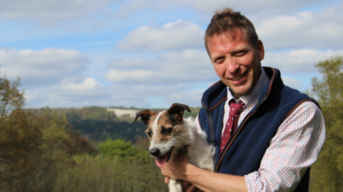 Yorkshire Vet Julian Norton - who is supporting elected mayor David Skaith's mens' mental health campaign - is smiling at the camera, holding Jack Russell dog Emmy and wearing a blue fleece waiscoat.