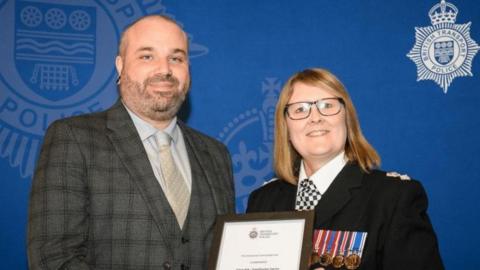 Simon Kirk accepting an award from a police officer with a blue backdrop 