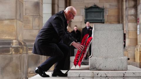 ohn Swinney crouches and lays a red poppy wreath at the Stone of Remembrance at the entrance to Edinburgh City Chambers on the Royal Mile. He has a black raincoat and highly polished black shoes.