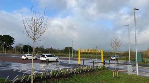 A car park with some cars scattered around in it. There is a yellow barrier at the entrance. It is a cloudy day and the ground is wet. 