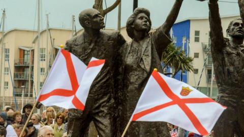 Jersey flags being waved in front of the island's liberation monument. The sculpture features two groups of symbolic islanders, including a child, plus a liberator. Crowds of people and buildings are in the background of the photo. 