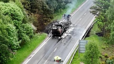 A smouldering burned-out lorry on one side of an empty road. Two firefighters are on the other side of the road