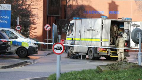 The bomb disposal squad at St James's Hospital in January 2023.  A large white van with a man opening the rear door dressed in camouflage style clothing and van has a yellow and blue line along the side and the words RC Bomb Disposal written on it in black - in front of the image is a line of police tape and behind the van is the red brick hospital building