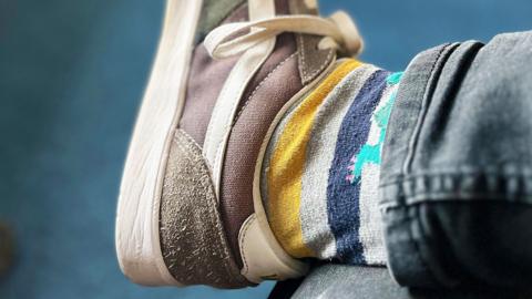 Close-up of a foot in a brown trainer, with stripey socks and blue jeans visible, against a blue background