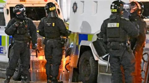 Two police Land Rovers in the background with four police officers in helmets standing in front of them. They are holding riot shields.