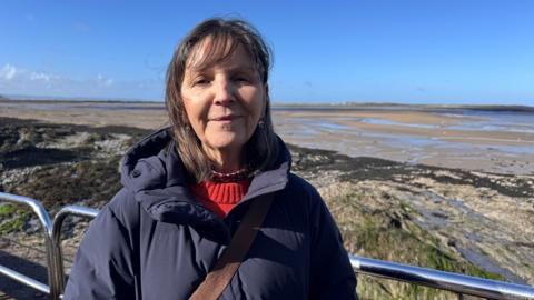 A woman with shoulder-length dark hair in a red jumper and thick navy coat stands in front of a wide beach, with rocks and sand behind her.