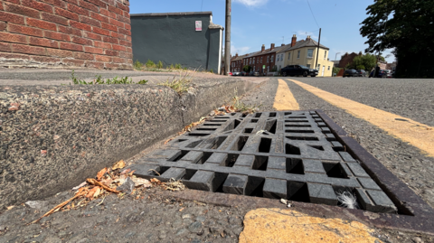 A drain cover in a road gutter, between double yellow lines. In the distance there's a row of houses.