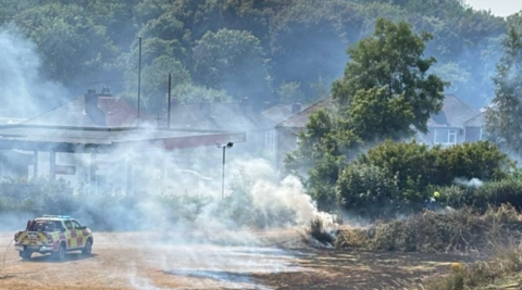 An emergency services vehicle is on the far left of the photo, which includes smoke from left to right. Houses and many trees are in the background.