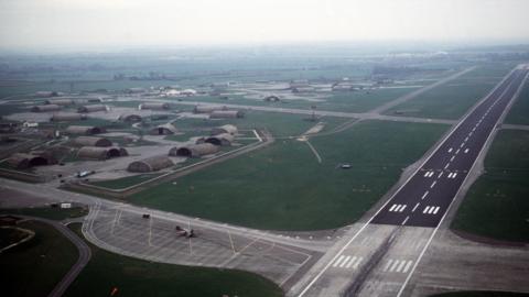 An aerial shot of a runway with green fields and low level buildings to the left.