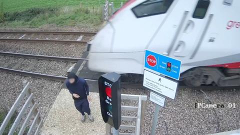 Masked toung person dressed in navy is stood with his back to a blurred train moving along the rails directly behind him.