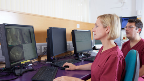 Two researchers in mauve overalls stare at a computer screen on a bank of desks.