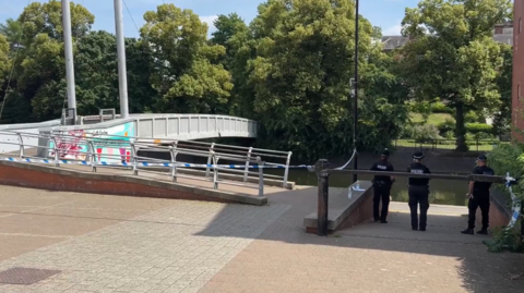 Three police officers stand by a taped-off area of river bank