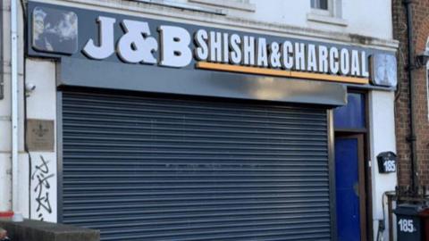 A shopfront with the words J&B Shisha & Charcoal printed in white on a black background above a closed metal roller shutter