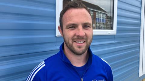 A man with dark hair and a beard standing in front of a light blue caravan. He is wearing a dark blue tracksuit top.