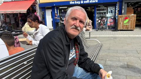 A man with a moustache and black Harrington jacket sits smiling on a bench while eating a sandwich.