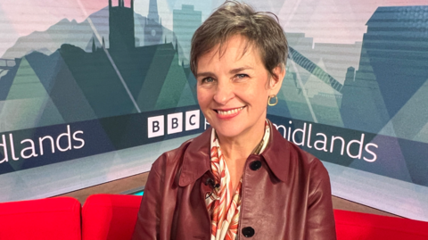 Mary Creagh MP has short brown hair, a red leather jacket and a red and white blouse with a floral pattern on a red sofa