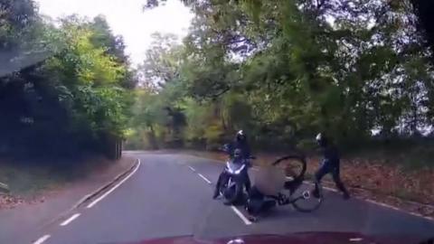A gang stand over a man on the ground who has been knocked off his bike on a country road.