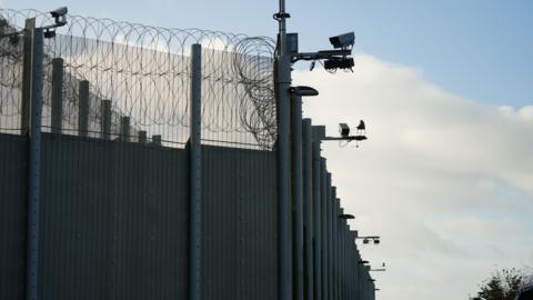 CCTV and barbed wire outside the La Moye prison fence which stands against a cloudy sky