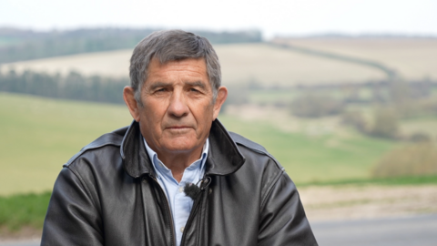 Phillip Wilkinson sits in front of hilly fields. He wears a leather jacket and a blue shirt. He has thick grey hair.