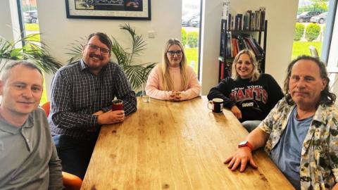 A group of five people sit around a large wooden table, drinking tea and coffee