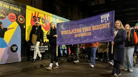 Two women holding a banner which reads Reclaim the Night walk along a street. there is a bright mural behind with the word Stop and some hands.