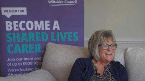 Kathy West is sitting on a grey sofa. She is laughing. Behind her is a poster saying: become a shared lives carer.