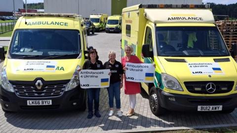 Three women are standing between two yellow ambulances that are park in front of large white buildings. Two of the women are holding up posters that state: "Ambulances 4 Ukraine." 