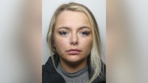 Katie Evans' mugshot from police. She has her hair up but some has fallen loose at the front. She has blond hair. She has eyelash extensions. She has freckles. She is wearing a grey top with a black puffer jacket.