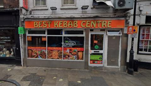 A view of the front of a kebab shop. A sign above the entrance reads Best Kebab Centre in gold letters against a red backdrop. 