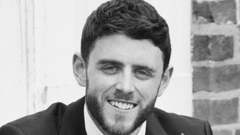 A black and white image of PC Andrew Harper a man with short brown hair and a slight beard wearing a suit and smiling at the camera.