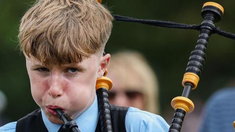 A close-up of a boy who is blowing his cheeks out as he plays on bagpipes. His eyes are half closed. 