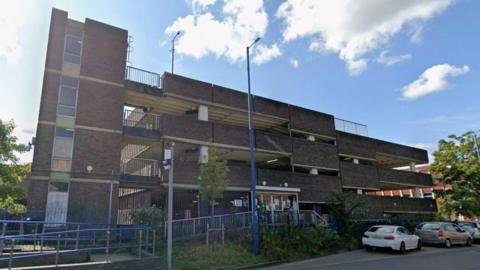 A view from the street of a Brutalist 1960s multi-storey car park, with four levels in red-grey brick 