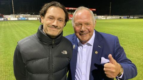 Frankie and Barry Fry standing on a floodlit pitch at Latimer Park. Frankie is wearing a black jumper with a black gilet over the top. He has long brown hair swept back and is smiling. Barry is wearing a light blue shirt with an unbuttoned navy blazer. He has short grey hair and is giving a thumbs up while smiling.