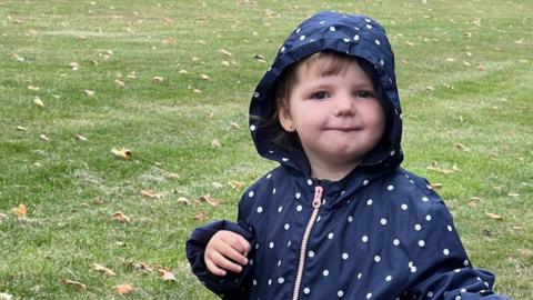 Maya, pictured standing in a park. She is wearing a blue hooded jacket with white dots. 