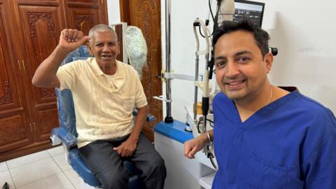 Indy Sian is sitting next to a patient in a treatment room. There is medical equipment behind him. Mr Sian is wearing blue scrubs and is smiling.